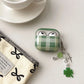 Green checkered airpod case with a shamrock charm on a white surface with a cup and saucer.
