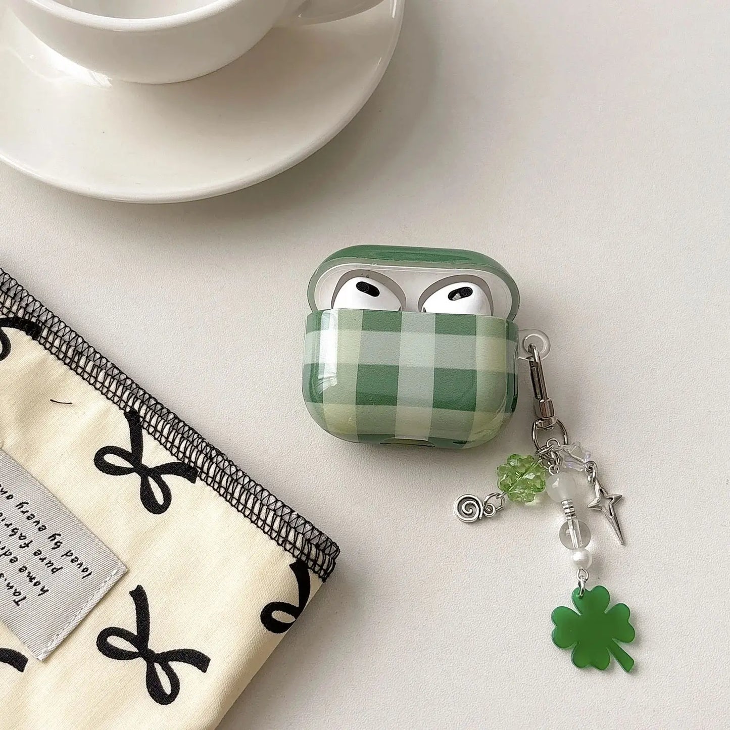 Green checkered airpod case with a shamrock charm on a white surface with a cup and saucer.
