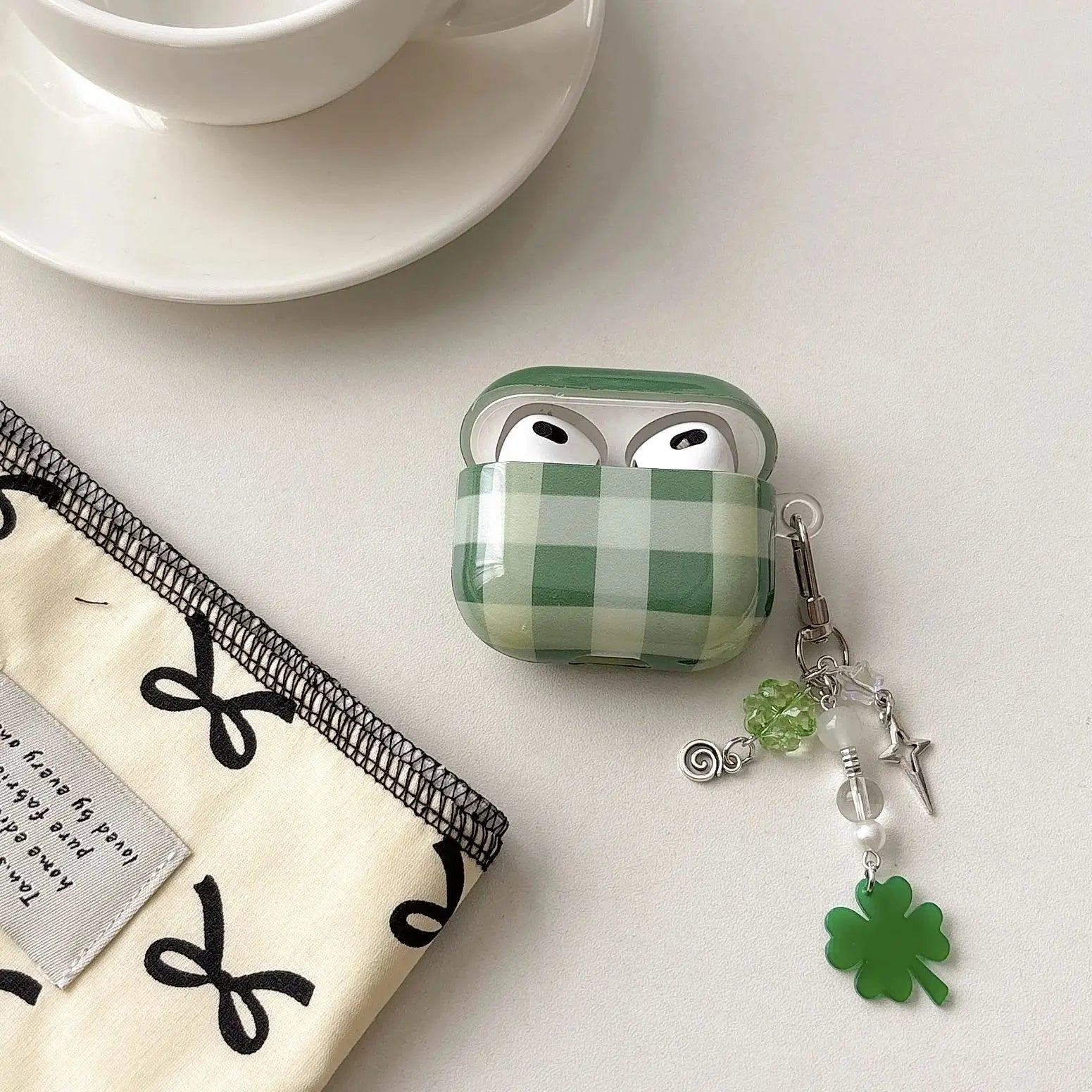 Green checkered airpod case with a shamrock charm on a white surface with a cup and saucer.