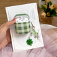 Green checkered AirPods case with white AirPods held by a hand on a white card, with a decorative keychain and flowers in the background.