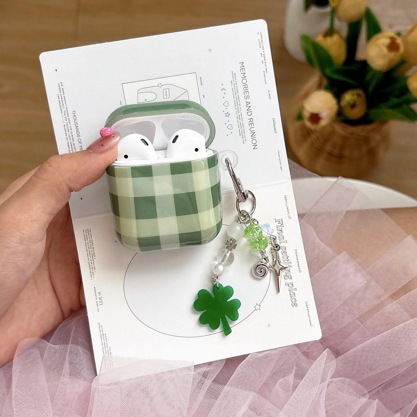 Green checkered AirPods case with white AirPods held by a hand on a white card, with a decorative keychain and flowers in the background.